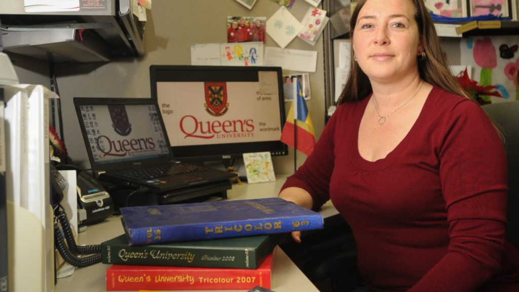 a USW female staff member sitting at a desk with mointors displaying the Queen's logo and a stack of tricolour Queen's yearbooks